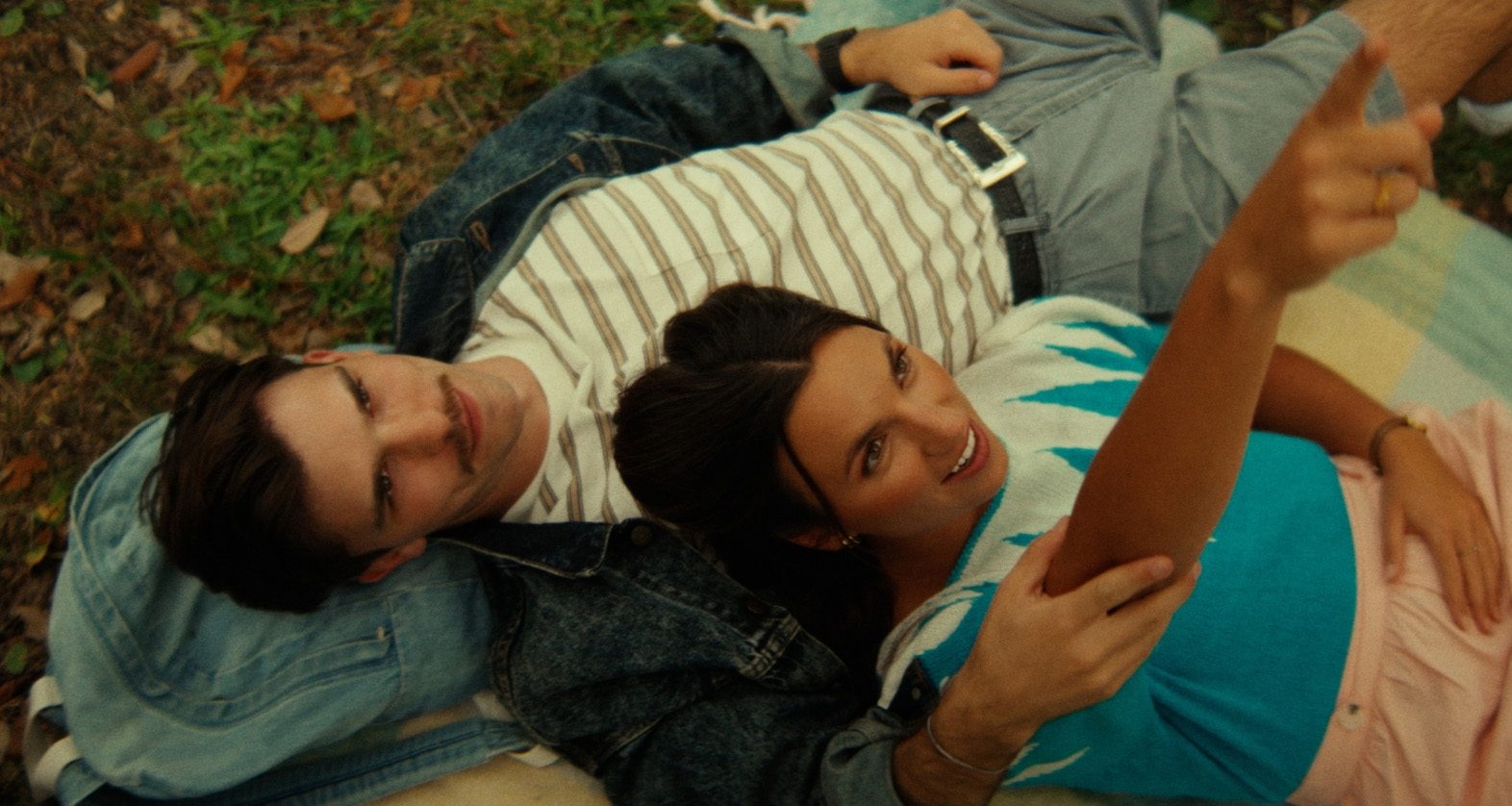 Overhead film still of a young couple lying head-to-head on a picnic blanket in a park, bathed in warm nostalgic light.